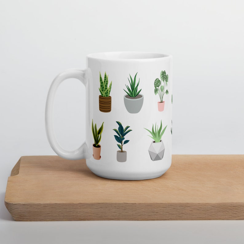May include: White ceramic mug featuring illustrations of various potted plants. The plants are depicted in different pots and styles, including snake plants, succulents, and a monstera. The mug is on a wooden surface.