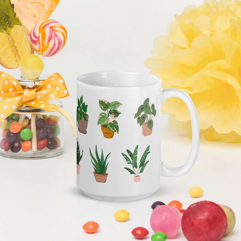 May include: White ceramic mug featuring illustrations of various potted plants. The plants include a variety of green and pink foliage. The mug has a large handle and is surrounded by colorful candies and a yellow decoration.