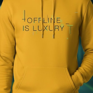 Puede incluir: Sudadera con capucha de color amarillo mostaza con el texto "OFFLINE IS LUXURY" en negro y verde. La sudadera tiene un bolsillo delantero y una capucha con cordón. El fondo es un degradado borroso de verde azulado y verde.