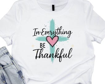 May include: White t-shirt with the message "In Everything Be Thankful" in black script. A teal cross and pink heart are centered on the shirt. Light blue denim jeans are folded at the bottom of the image.
