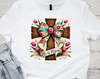 May include: White t-shirt with a brown wooden cross with a floral bow and the text "He is Risen" on a banner.