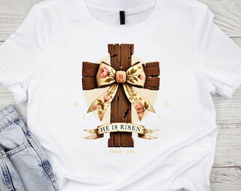 May include: White t-shirt with a brown wooden cross with a floral bow and the text "He is Risen" on a banner.