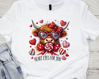 Pode incluir: Camiseta branca com uma vaca Highland de desenho animado usando óculos de sol em forma de coração e um pirulito. A vaca tem um laço vermelho e é cercada por decorações em forma de coração. O texto "HEART EYES FOR YOU" está impresso abaixo.