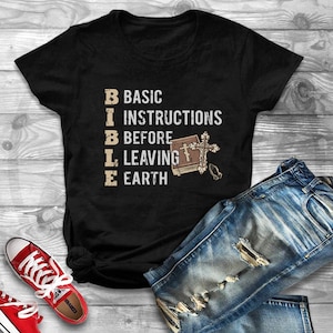 May include: Black t-shirt with white text that reads "BIBLE: BASIC INSTRUCTIONS BEFORE LEAVING EARTH". The text is arranged vertically with a small image of a bible and cross next to it.