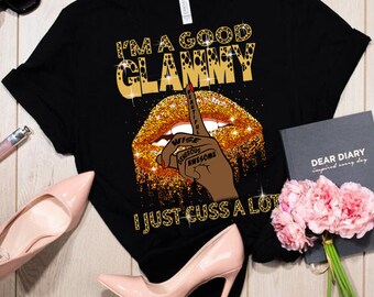Op de afbeelding: Zwart T-shirt met gouden tekst "I'M A GOOD GLAMMY I JUST CUSS A LOT." Het ontwerp bevat een gouden glitter lip-afbeelding met een vinger naar de lippen gebaar. De vinger heeft de woorden "BEAUTIFUL", "WISE", "GENEROUS" en "AWESOME".