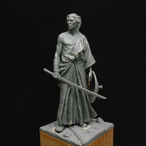 May include: A gray painted figurine of a samurai warrior holding a sword and a shield. The figurine is standing on a wooden base.