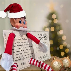 Peut inclure: Une poupée elfe de Noël avec un bonnet de Père Noël rouge et blanc et des jambes rayées rouges et blanches. L'elfe tient une carte postale avec du texte en français. Des boules de Noël dorées sont au premier plan.