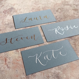 May include: Four place cards with names written in calligraphy. The cards are a light blue colour and the names are written in gold and white ink.