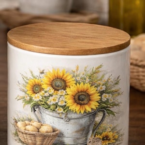 May include: A white ceramic canister with a wooden lid, featuring a painted design of sunflowers and daisies in a metal bucket. A small basket of potatoes is also depicted. The canister is a kitchen storage item.
