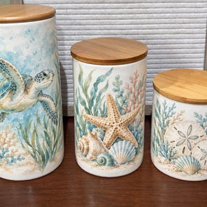 May include: A set of three cylindrical ceramic canisters with bamboo lids. The tallest features a sea turtle, the medium a starfish and seashells, and the shortest displays sand dollars and seashells, all with watercolour-style aquatic plant designs.