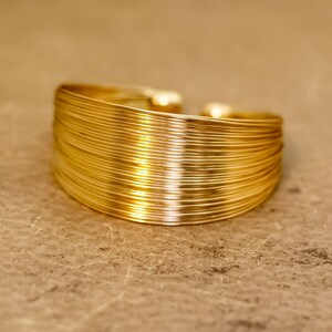Puede incluir: Un brazalete dorado hecho de alambres finos entrelazados. El brazalete es curvo y tiene un acabado liso y pulido.