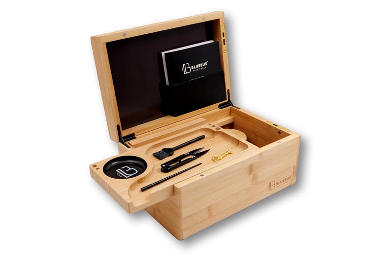 K&ouml;nnte beinhalten: Eine helle Bambusbox mit offenem Deckel, die einen schwarzen Innenraum und ein Tablett mit Werkzeugen freigibt. Das Tablett enth&auml;lt eine B&uuml;rste, eine Schere, einen Poker und ein kleines goldfarbenes Werkzeug. Ein kleiner schwarzer Beh&auml;lter mit dem "B"-Logo befindet sich links. Die Box hat das "BLUEBUS"-Logo.