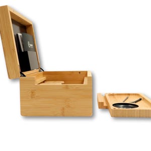K&ouml;nnte beinhalten: Eine Bambusbox mit Scharnierdeckel, die sich &ouml;ffnet und ein Staufach und einen schwarzen Kartenhalter freigibt. Ein separates Bambustablett mit einer kreisf&ouml;rmigen schwarzen Schale und Werkzeugen befindet sich neben der Box.