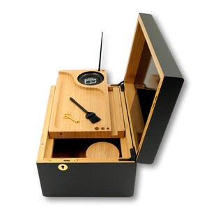 K&ouml;nnte beinhalten: Eine schwarze und nat&uuml;rliche Holzkiste mit offenem Deckel, die ein Holztablett mit einer kleinen B&uuml;rste, einem Schl&uuml;ssel und einem kreisf&ouml;rmigen Beh&auml;lter freilegt. Die Box hat ein Schloss und eine kleine Antenne. Der Innenraum ist aus Naturholz und der Au&szlig;enbereich ist schwarz.