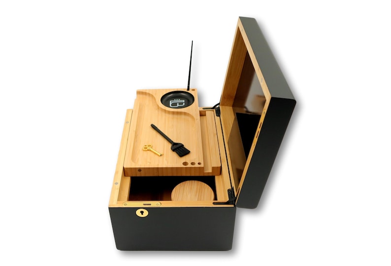 K&ouml;nnte beinhalten: Eine schwarze und nat&uuml;rliche Holzkiste mit offenem Deckel, die ein Holztablett mit einer kleinen B&uuml;rste, einem Schl&uuml;ssel und einem kreisf&ouml;rmigen Beh&auml;lter freilegt. Die Box hat ein Schloss und eine kleine Antenne. Der Innenraum ist aus Naturholz und der Au&szlig;enbereich ist schwarz.