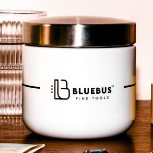 May include: A white container with a silver lid and the text "BLUEBUS FINE TOOLS" in black. The container is cylindrical and sits on a wooden surface. A clear glass and a yellow container are in the background.