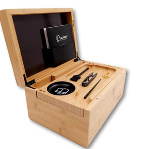 May include: A light-colored bamboo box with an open lid, revealing various tools and accessories. The box contains a black bowl, a brush, scissors, a poker, and other items. A black container with white text is inside the box.