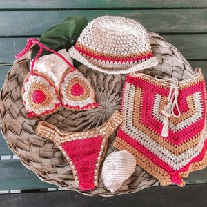 May include: A crocheted bikini set in pink, beige, and white with a matching crocheted hat. The bikini top is a triangle style with a floral design. The bikini bottom is a high-waisted style with a scalloped edge. The hat is a bucket style with a wide brim.