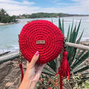 May include: Red crocheted round crossbody bag with a gold chain strap and a red tassel. The bag has a wooden bead accent and a brown leather tag that says "SOUL TAY".
