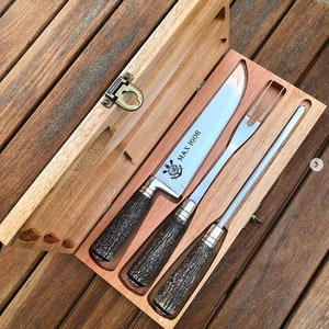 Könnte beinhalten: Eine Holzbox mit einem Tranchiermesser, einer Gabel und einem Wetzstahl. Messer und Gabel haben dunkelbraune, strukturierte Griffe und silberne Klingen. Die Messerklinge trägt den Schriftzug "MAX IGOR" und ein nautisches Emblem.