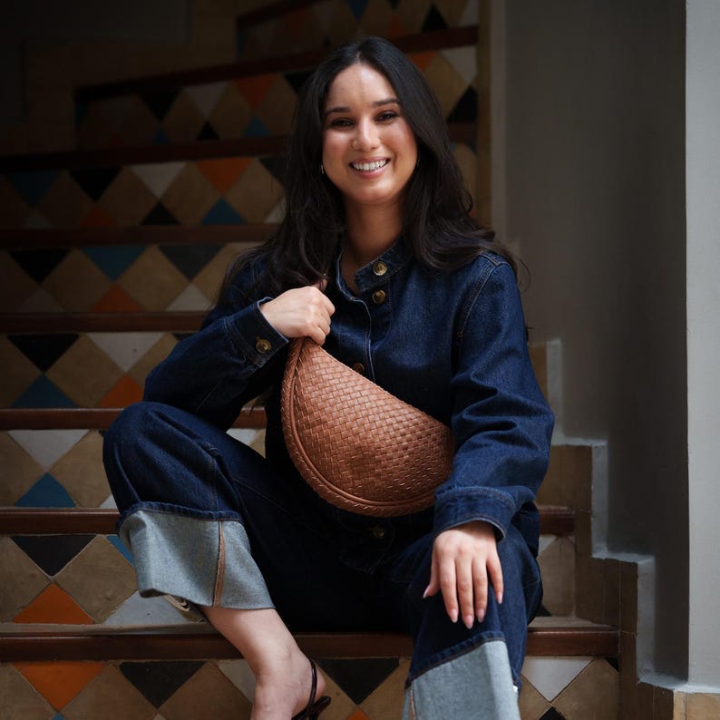 May include: A woman wearing a denim jacket and jeans, sitting on a staircase. She is holding a woven, tan-colored, half-moon shaped bag. The staircase has a decorative tile pattern in shades of brown, blue, and orange.