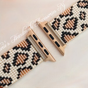 Puede incluir: Una banda de reloj Apple de tono dorado con un patrón de cuentas en blanco, negro y marrón. La banda está hecha de pequeñas cuentas tejidas juntas para crear un diseño de estampado de leopardo.
