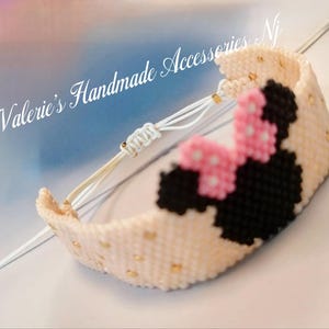 Pode incluir: Um bracelete de amizade branco e dourado com um design de Minnie Mouse preto e rosa. O bracelete é feito com pequenas contas e tem um fecho de corda branco.