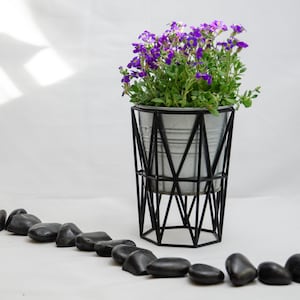 May include: A metal planter with a geometric black stand, filled with purple flowers and sitting on a white surface with black stones.