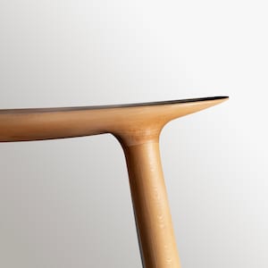 Può includere: Primo piano di una gamba di tavolo in legno chiaro con una parte superiore curva. La gamba del tavolo è realizzata in legno massello e ha una finitura naturale.