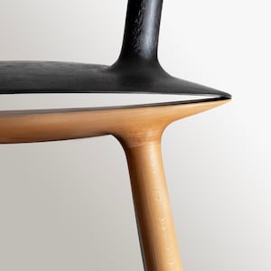 Op de afbeelding: Close-up van een houten stoel met een zwart gebeitst zitting en een natuurlijke houten basis. De stoel heeft een uniek ontwerp met een gebogen zitting en poten.
