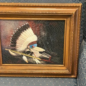 May include: Framed artwork featuring a Native American headdress with white and black feathers, blue and red accents, and a skull. The painting has a dark background with a cracked texture and is set in a gold-toned wooden frame.