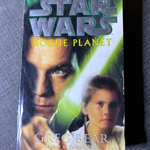 May include: A paperback book cover for the Star Wars novel "Rogue Planet" by Greg Bear. The cover features a green and white background with a lightsaber beam and two characters, one with a dark complexion and the other with a light complexion.