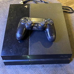 Op de afbeelding: Een zwarte PlayStation 4 console met bijpassende controller. De console is rechthoekig met een matte afwerking aan de rechterkant en een glanzende afwerking aan de linkerkant. De controller ligt erop. De console is ongeveer 30 cm lang.