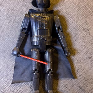 May include: A black Darth Vader action figure with a red lightsaber. The figure has a black cape and is standing on a gray surface. The figure is a vintage collectible.