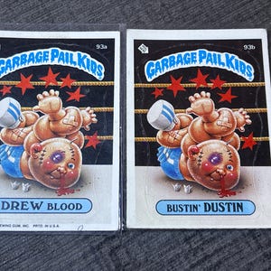Op de afbeelding: Twee Garbage Pail Kids-verzamelkaarten, "Drew Blood" en "Bustin' Dustin". Elke kaart toont een stripfiguur met een blauw oog en verbanden, in een worstelringomgeving. De kaarten hebben het Garbage Pail Kids-logo.