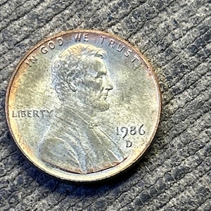 May include: A copper-colored United States one-cent coin featuring the profile of Abraham Lincoln facing right. The coin is inscribed with the words IN GOD WE TRUST, LIBERTY, and the year 1986 with a D mint mark below the date.