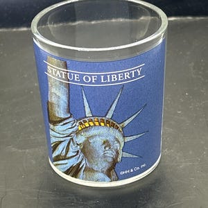 Könnte beinhalten: Ein klares Schnapsglas mit blauem Hintergrund, das eine Illustration der Freiheitsstatue zeigt. Der Text "STATUE OF LIBERTY" ist über dem Bild aufgedruckt. Auf dem Boden des Glases steht "©HH & Co. Inc."