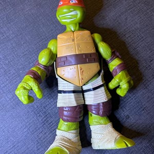 May include: A green action figure of a Teenage Mutant Ninja Turtle, wearing an orange mask. The figure has a tan shell, brown belt, and white and brown wraps on its legs and arms. The figure is standing on a gray surface.
