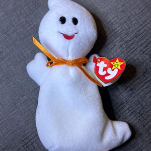 Puede incluir: Un juguete de fantasma de peluche blanco con ojos negros y una boca roja sonriente. Tiene una cinta naranja atada alrededor del cuello y una etiqueta en forma de corazón. La etiqueta dice "TY Beanie Babies" y "Spooky".