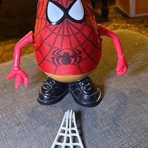 Può includere: Un giocattolo di Mr. Potato Head a tema Spider-Man con un design a ragnatela rosso e nero sulla testa e sul busto. Il giocattolo ha scarpe nere, braccia rosa e un accessorio a ragnatela bianca. Un giocattolo da collezione divertente.