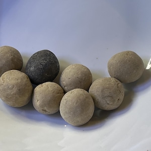 Pode incluir: Uma coleção de oito objetos esféricos, maioritariamente bege, dispostos numa tigela branca. Um objeto é cinzento mais escuro. Os objetos parecem ser feitos de um material natural. A tigela é branca e tem um design simples e limpo.