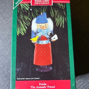 May include: A Hallmark Keepsake Ornament featuring a red and white nutcracker doll with a blue hat and a yellow bird in its hand. The ornament is labeled "Frieda The Animals' Friend" and "North Pole Nutcrackers".