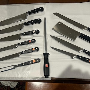 May include: A collection of kitchen knives with black handles and stainless steel blades. Includes a carving fork, a cleaver, a bread knife, and various chef's knives. A sharpening steel is also present.