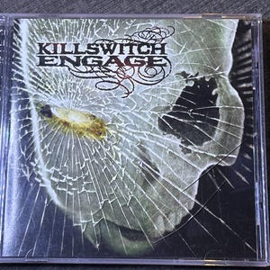 Può includere: Un'immagine della custodia del CD dell'album "As Daylight Dies" dei Killswitch Engage. La copertina presenta un'immagine in scala di grigi incrinata con il nome della band in grassetto nero e dettagli rossi.