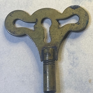 May include: Antique brass clock key with a decorative top and a keyhole shape. The key has a cylindrical stem and a tarnished finish, showing signs of age and use. A vintage clock part.