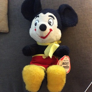 May include: A vintage plush Mickey Mouse toy with black ears, a white face, red shorts, and yellow shoes. The toy has a yellow ribbon around its neck and a tag that reads "Walt Disney's Mickey Mouse".