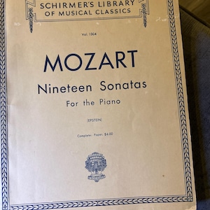 May include: A vintage music book titled "MOZART Nineteen Sonatas For the Piano" by Schirmer's Library of Musical Classics. The cover is beige with blue text and a decorative border. The book is a paper edition.