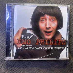 Puede incluir: Una caja de disco compacto con una portada que muestra un retrato de Emo Philips. El texto dice E=MO2, EMO PHILIPS y PLUS THE ENTIRE LIVE AT THE HASTY PUDDING THEATRE. El texto del lomo lateral dice TWO CLASSIC EMO PHILIPS ALBUMS ON ONE CD.