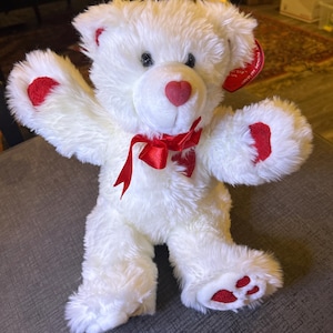 Könnte beinhalten: Ein weißer Teddybär mit roten Akzenten an Ohren, Pfoten und Nase. Der Bär hat eine rote herzförmige Nase und eine rote Schleife. Ein herzförmiges Etikett ist am Ohr befestigt.