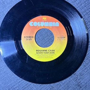 May include: A black vinyl record with a yellow and orange label. The label features the Columbia Records logo and the text "ROSANNE CASH SEVEN YEAR ACHE". The record is a 45 RPM record and was released in 1981.